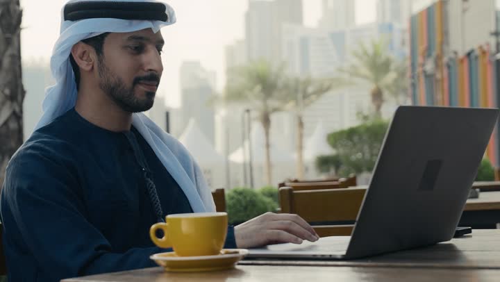 الاستمتاع بشرب القهوه ، إنجاز مهام العمل عن بعد، إدارة شؤون الأعمال وتنظيمها ، استخدام التكنولوجيا والتقنيات المتطورة، تصوير لرجل عربي خليجي اماراتي يرتدي الكندورة والغترة يجلس على كرسي في إحدى المقاهي يشرب مشروب ساخن ويستخدم الحاسوب المحمول .