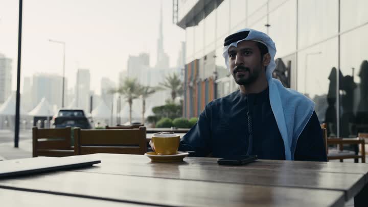 إنجاز مهام العمل عن بعد، إدارة شؤون الأعمال وتنظيمها ، الاستمتاع بشرب القهوه ، استخدام التكنولوجيا والتقنيات المتطورة، تصوير لرجل عربي خليجي اماراتي يرتدي الكندورة والغترة يجلس على كرسي في إحدى المقاهي يشرب مشروب ساخن ويستخدم الحاسوب المحمول .