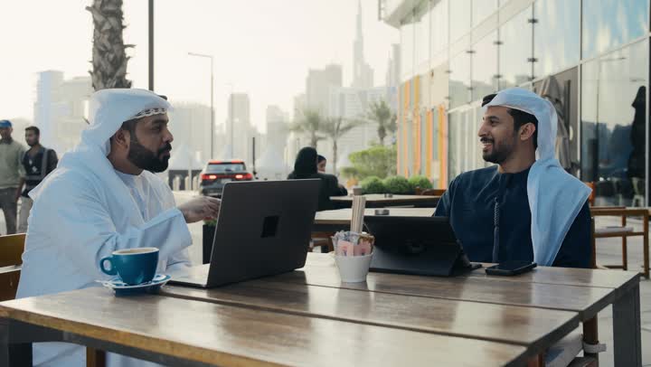 مفهوم التكنولوجيا وإستخدام أجهزة تقنية حديثة ، قضاء وقت ممتع مع الأصدقاء, استخدم جهاز الحاسوب المحمول لانجاز المشاريع والمهام في الكافيه ،  تصوير مقرب لشاب عربي خليجي إماراتي يرتدي الكندورة والغترة البيضاء يجلس مع زميل عمل في المقهى .