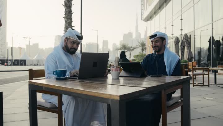 Dialogue and discussion between colleagues, spending quality time with friends, using a laptop to complete projects and tasks in the café, the concept of technology and the use of modern technical devices, a close-up of an Emirati Gulf Arab young man wearing a white kandura and ghutra sitting with a colleague in the café.