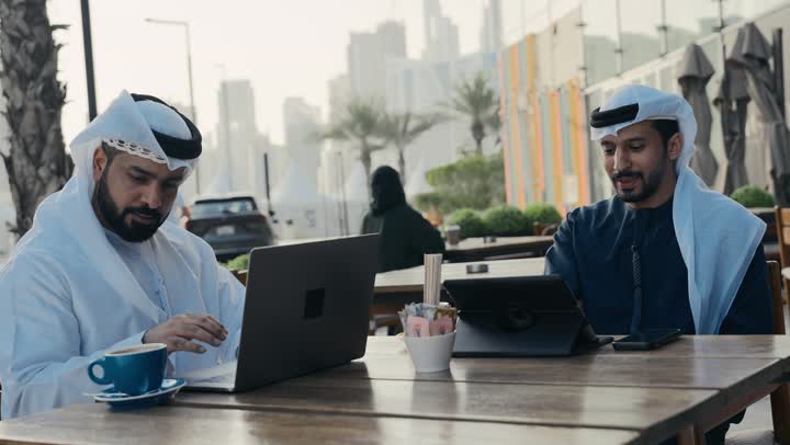 قضاء وقت ممتع مع الأصدقاء, استخدم جهاز الحاسوب المحمول لانجاز المشاريع والمهام في الكافيه ، مفهوم التكنولوجيا وإستخدام أجهزة تقنية حديثة ، تصوير مقرب لشاب عربي خليجي إماراتي يرتدي الكندورة والغترة البيضاء يجلس مع زميل عمل في المقهى .