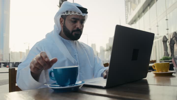 إدارة شؤون الأعمال وتنظيمها ، الاستمتاع بشرب القهوه ، إنجاز مهام العمل عن بعد، استخدام التكنولوجيا والتقنيات المتطورة، تصوير لرجل عربي خليجي اماراتي يرتدي الكندورة والغترة يجلس على كرسي في إحدى المقاهي يشرب مشروب ساخن ويستخدم الحاسوب المحمول .
