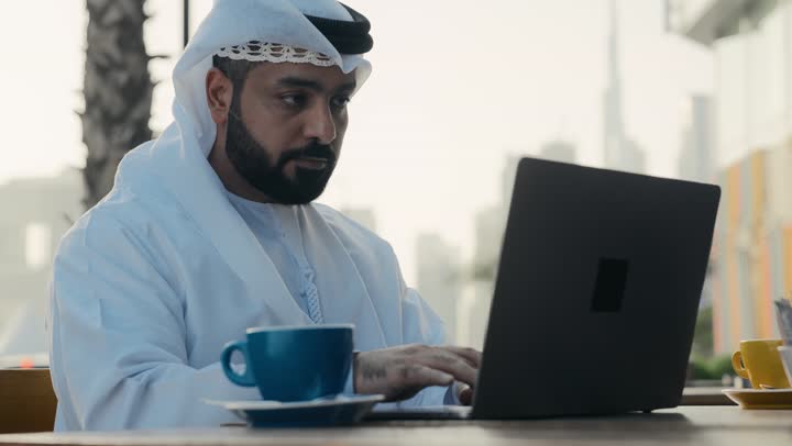 الاستمتاع بشرب القهوه ، إنجاز مهام العمل عن بعد، استخدام التكنولوجيا والتقنيات المتطورة، إدارة شؤون الأعمال وتنظيمها عبر الأجهزة التقنية الحديثة، تصوير لرجل عربي خليجي اماراتي يرتدي الكندورة والغترة البيضاء يجلس على كرسي في إحدى المقاهي يشرب مشروب ساخن ويستخدم الحاسوب المحمول .