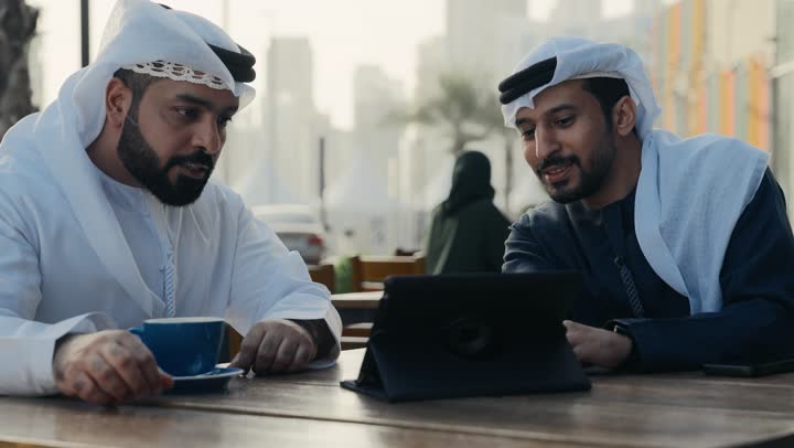 استخدم جهاز الحاسوب المحمول لانجاز المشاريع والمهام في الكافيه ، مفهوم التكنولوجيا وإستخدام أجهزة تقنية حديثة ، قضاء وقت ممتع مع الأصدقاء, تصوير مقرب لشاب عربي خليجي إماراتي يرتدي الكندورة والغترة البيضاء يجلس مع زميل عمل في المقهى .