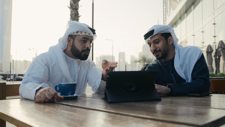 The concept of technology and the use of modern technological devices, using a laptop to complete projects and tasks in the café, spending quality time with friends, a close-up of an Emirati Gulf Arab young man wearing a kandura and a white ghutra sitting with a colleague in the café.