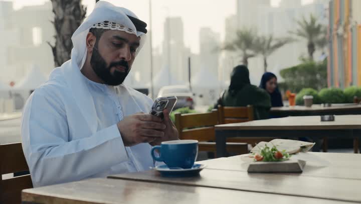 التسلية والترفيه في العطلة، تصفح مواقع التواصل الاجتماعي عبر الانترنت ، الاستمتاع بالاجواء الهادئة خارج المنزل، تصوير لرجل عربي خليجي اماراتي يرتدي الكندورة والغترة البيضاء يجلس على كرسي في إحدى المقاهي الراقية يمسك الهاتف المحمول