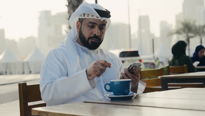 تصفح مواقع التواصل الاجتماعي عبر الانترنت ، الاستمتاع بالاجواء الهادئة خارج المنزل، تصوير لرجل عربي خليجي اماراتي يرتدي الكندورة والغترة البيضاء يجلس على كرسي في إحدى المقاهي الراقية يمسك الهاتف المحمول ،التسلية والترفيه في العطلة