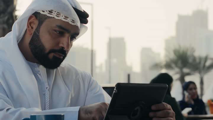 إنجاز مهام العمل عن بعد، استخدام التكنولوجيا والتقنيات المتطورة، إدارة شؤون الأعمال وتنظيمها عبر الأجهزة التقنية الحديثة، تصوير لرجل عربي خليجي اماراتي يرتدي الكندورة والغترة البيضاء يجلس على كرسي في إحدى المقاهي يشرب مشروب ساخن ويستخدم الحاسوب المحمول .