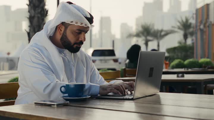 استخدام التكنولوجيا والتقنيات المتطورة،إدارة شؤون الأعمال وتنظيمها عبر الأجهزة التقنية الحديثة، إنجاز مهام العمل عن بعد، تصوير لرجل عربي خليجي اماراتي يرتدي الكندورة والغترة البيضاء يجلس على كرسي في إحدى المقاهي يشرب مشروب ساخن ويستخدم الحاسوب المحمول .