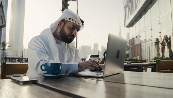 Managing and organizing business affairs using modern technical devices, using advanced technology and techniques, completing work tasks remotely. A picture of an Emirati Gulf Arab man wearing a white kandura and ghutra sitting on a chair in a cafe, drinking a hot drink and using a laptop.