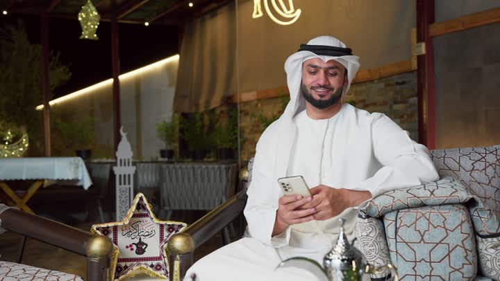 Ramadan hospitality, expressions of joy and happiness, using a modern technical device, a young Emirati Gulf Arab man wearing a kandura and ghutra using a mobile phone sitting outdoors, Ramadan decorations and lighting, an outdoor evening session in one of the chalets in the Emirates.
