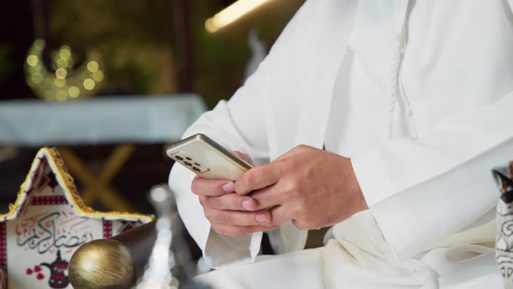 تعابير السرور والسعادة، ضيافة شهر رمضان المبارك، استخدام جهاز تقني حديث، شاب عربي خليجي اماراتي يرتدي الكندورة والغترة يستخدم الجوال يجلس في الهواء الطلق، زينة واضاءة رمضانية، الجلسة الليلة الخارجية في احدى شاليهات الامارات