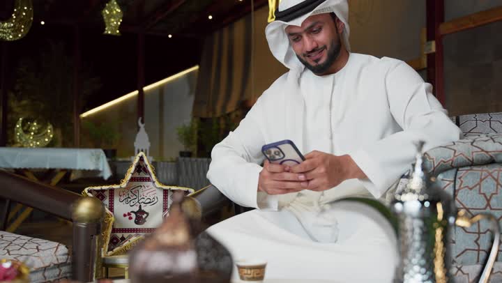 An outdoor evening session in one of the chalets in the Emirates, modern technology and techniques, expressions of pleasure and happiness, hospitality of the holy month of Ramadan, a young Emirati Gulf Arab man wearing a kandura and a ghutra using a mobile phone sitting outdoors, Ramadan decorations and lighting, using a modern device