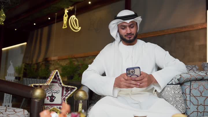 Hospitality of the holy month of Ramadan, an outdoor evening session in one of the chalets in the Emirates, modern technology and techniques, expressions of pleasure and happiness, a young Emirati Gulf Arab man wearing a kandura and a ghutra using a mobile phone sitting in the open air