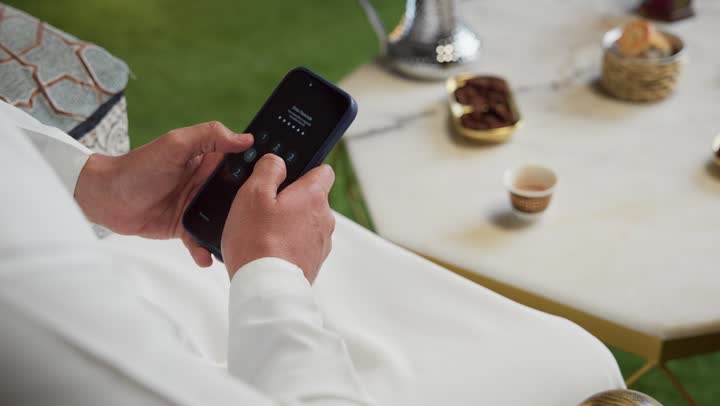 Integrating technology into daily life, Ramadan hospitality, an outdoor evening session in one of the UAE chalets, modern technology and techniques, expressions of pleasure and happiness, a young Emirati Gulf Arab man wearing a kandura and ghutra using a mobile phone sitting outdoors.