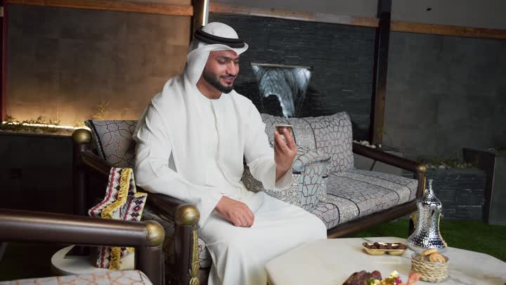 اجراء بث مباشر ، دمج التكنولوجيا فى الحياه اليوميه، ضيافة شهر رمضان المبارك،جلسة الليلة الخارجية في احدى شاليهات الامارات ، تكنولوجيا وتقنيات حديثه ،شاب عربي خليجي اماراتي يرتدي الكندورة والغترة يستخدم الجوال يجلس في الهواء الطلق