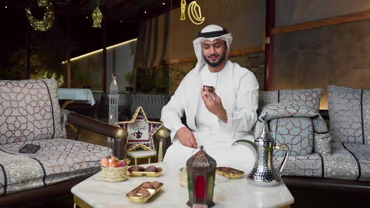 Making video calls, integrating technology into daily life, hosting the holy month of Ramadan, an outdoor evening session in one of the chalets in the Emirates, modern technology and techniques, a young Emirati Gulf Arab man wearing a kandura and ghutra using a mobile phone sitting outdoors.