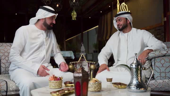 Two Emirati Gulf Arab friends wearing kandura and ghutra sitting outdoors, warm welcome and generosity in the holy month of Ramadan, traditional coffee drinking customs and traditions, breathing fresh air, enjoying the happy Ramadan atmosphere, spending quality time with friends, outdoor evening session in one of the chalets in the Emirates