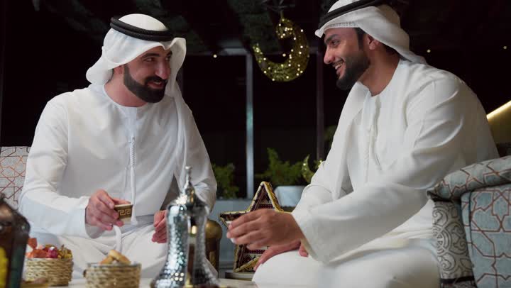 قضاء اوقات ممتعه مع الاصدقاء ، الجلسة الليلة الخارجية في احدى شاليهات الامارات، عادات وتقاليد شرب القهوة التقليدية، حسن الاستقبال والكرم في شهر رمضان المبارك، صديقان عربيان خليجيان اماراتيان يرتديان الكندورة والغترة يجلسان في الهواء الطلق، زينة واضاءة رمضانية