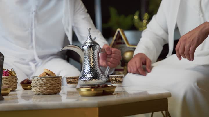 الجلسة الليلة الخارجية في احدى شاليهات الامارات، عادات وتقاليد شرب القهوة التقليدية، حسن الاستقبال والكرم في شهر رمضان المبارك، صديقان عربيان خليجيان اماراتيان يرتديان الكندورة والغترة يجلسان في الهواء الطلق، منتج وطني محلي اماراتي، زينة واضاءة رمضانية