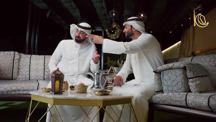 Setting a specific goal, an outdoor evening session in one of the chalets in the Emirates, the customs and traditions of drinking traditional coffee, good reception and generosity in the holy month of Ramadan, two Emirati Gulf Arab friends wearing the kandura and ghutra sitting outdoors, a local Emirati national product, Ramadan decorations and lighting
