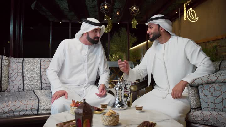 عادات وتقاليد شرب القهوة التقليدية، الجلسة الليلة الخارجية في احدى شاليهات الامارات، حسن الاستقبال والكرم في شهر رمضان المبارك، صديقان عربيان خليجيان اماراتيان يرتديان الكندورة والغترة يجلسان في الهواء الطلق، منتج وطني محلي اماراتي، زينة واضاءة رمضانية
