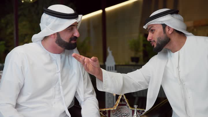الاشاره الى شئ ما ، الجلسة الليلة الخارجية في احدى شاليهات الامارات، عادات وتقاليد شرب القهوة التقليدية، حسن الاستقبال والكرم في شهر رمضان المبارك، صديقان عربيان خليجيان اماراتيان يرتديان الكندورة والغترة يجلسان في الهواء الطلق، منتج وطني محلي اماراتي، زينة واضاءة رمضانية