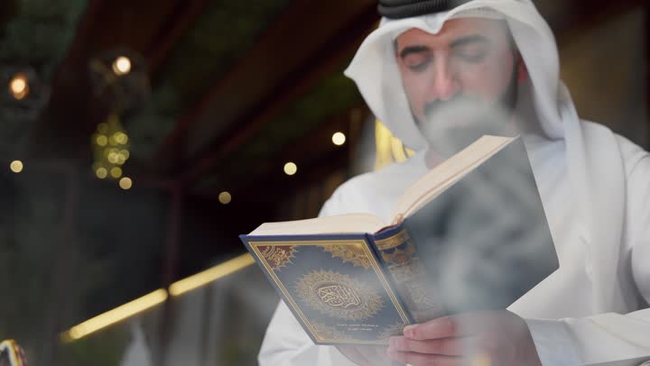الإكثار من ذكر الله تعالى، التقرب الى الله في شهر رمضان، الجلوس فى احدي الشاليهات الاماراتيه ، تلاوة وقراءة القرآن الكريم وتدبر معانيه، الشعور بالخشوع والطمأنينة، الاجتهاد في العبادة، شاب عربي خليجي اماراتي يرتدي الكندوره والغتره يحمل بيده المصحف الشريف
