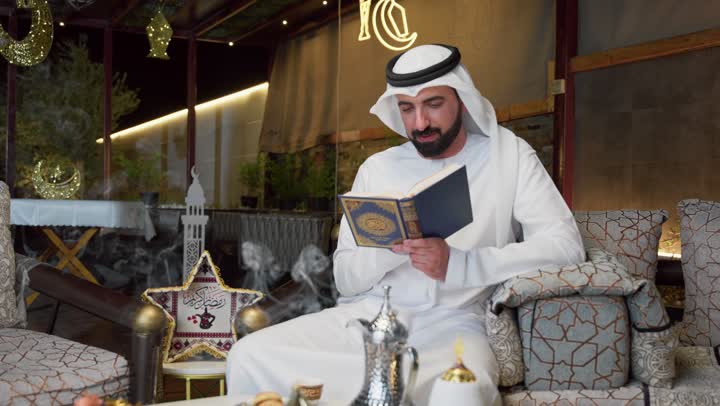 الاجتهاد في العبادة ، شاب عربي خليجي اماراتي يرتدي الكندوره والغتره يحمل بيده المصحف الشريف ، الإكثار من ذكر الله تعالى، التقرب الى الله في شهر رمضان، الجلوس فى احدي الشاليهات الاماراتيه ، تلاوة وقراءة القرآن الكريم وتدبر معانيه، الشعور بالخشوع والطمأنينة