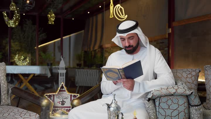 شاب عربي خليجي اماراتي يرتدي الكندوره والغتره يحمل بيده المصحف الشريف ، الإكثار من ذكر الله تعالى، التقرب الى الله في شهر رمضان، الجلوس فى احدي الشاليهات الاماراتيه ، تلاوة وقراءة القرآن الكريم وتدبر معانيه، الشعور بالخشوع والطمأنينة، الاجتهاد في العبادة