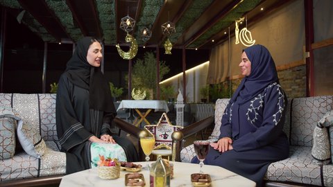 تبادل الاحاديث والاخبار ، زينه وديكورات رمضانيه ، حسن الاستقبال وكرم الضيافة في شهر رمضان، جلسة ليلية خارجية في إحدى شاليهات الامارات، قضاء وقت ممتع مع الأصدقاء ، الاستمتاع بشرب المشروبات البارده ،صديقتان عربيتان خليجيتان اماراتيان ترتديان العباءة والحجاب تجلسان في الهواء الطلق بايماءات السعاده