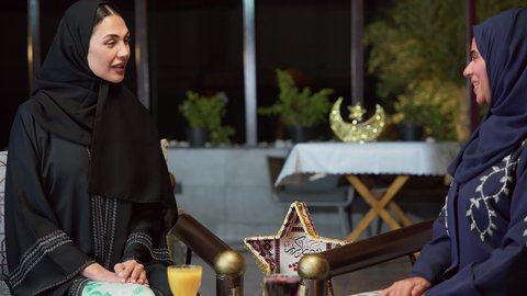 تبادل الاحاديث والاخبار ، زينه وديكورات رمضانيه ، حسن الاستقبال وكرم الضيافة في شهر رمضان، جلسة ليلية خارجية في إحدى شاليهات الامارات، قضاء وقت ممتع مع الأصدقاء ، الاستمتاع بشرب المشروبات البارده ،صديقتان عربيتان خليجيتان اماراتيان ترتديان العباءة والحجاب تجلسان في الهواء الطلق بايماءات السعاده