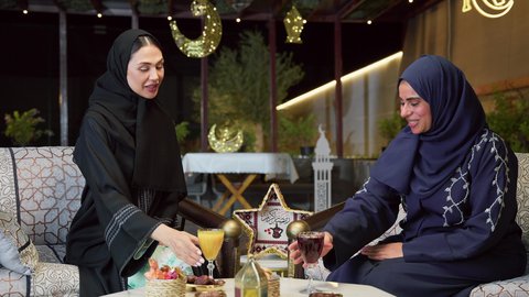 جلسة ليلية خارجية في إحدى شاليهات الامارات، قضاء وقت ممتع مع الأصدقاء ، الاستمتاع بشرب المشروبات البارده ،صديقتان عربيتان خليجيتان اماراتيان ترتديان العباءة والحجاب تجلسان في الهواء الطلق يتبادلان الاحاديث والاخبار بتعابير السرور، حسن الاستقبال وكرم الضيافة في شهر رمضان