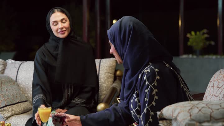 قضاء وقت ممتع مع الأصدقاء ، الاستمتاع بشرب المشروبات البارده ،صديقتان عربيتان خليجيتان اماراتيان ترتديان العباءة والحجاب تجلسان في الهواء الطلق يتبادلان الاحاديث والاخبار بتعابير السرور، حسن الاستقبال وكرم الضيافة في شهر رمضان، جلسة ليلية خارجية في إحدى شاليهات الامارات