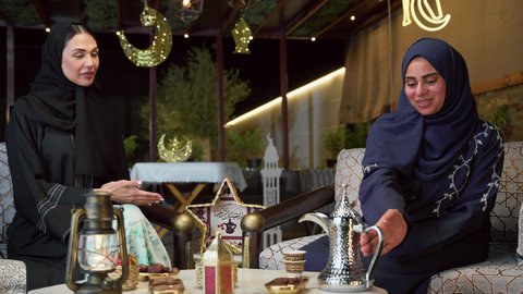 قضاء وقت ممتع مع الأصدقاء ، صب القهوه العربيه، حسن الاستقبال وكرم الضيافة في شهر رمضان، جلسة ليلية خارجية في إحدى شاليهات الامارات، الاستمتاع بشرب المشروبات الساخنه ، صديقتان عربيتان خليجيتان اماراتيان ترتديان العباءة والحجاب تجلسان في الهواء الطلق يتبادلان الاحاديث والاخبار بتعابير السرور