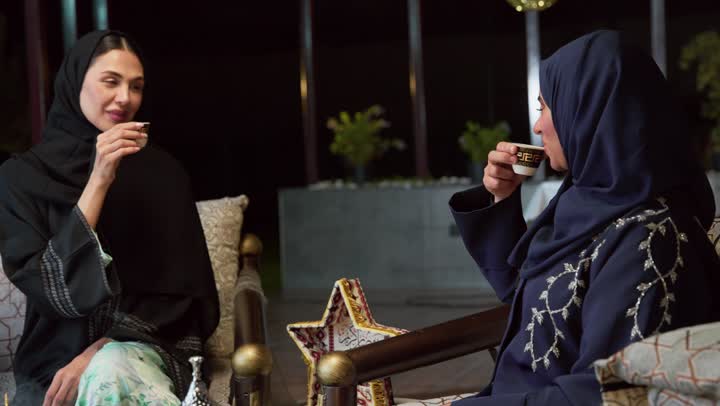 Enjoying hot drinks, two Emirati Gulf Arab friends wearing abaya and hijab sitting outdoors exchanging conversations and news with happy expressions, an outdoor night session in one of the chalets in the Emirates, warm welcome and hospitality in the month of Ramadan, spending a pleasant time with friends