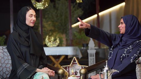 تحديد هدف معين ، صديقتان عربيتان خليجيتان اماراتيان ترتديان العباءة والحجاب تجلسان في الهواء الطلق يتبادلان الاحاديث والاخبار بتعابير السرور، جلسة ليلية خارجية في إحدى شاليهات الامارات، حسن الاستقبال وكرم الضيافة في شهر رمضان، قضاء وقت ممتع مع الأصدقاء