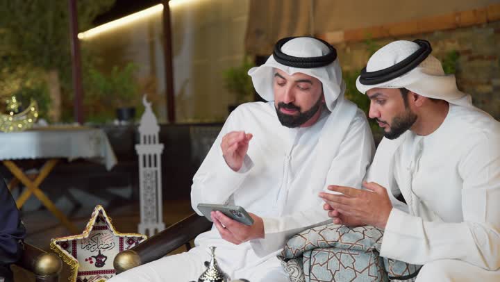 Using modern technology and techniques, two Emirati Gulf Arab friends wearing kandura and ghutra sitting outdoors exchanging conversations and news with happy expressions, an outdoor night session in one of the chalets in the Emirates, warm reception and hospitality in the month of Ramadan, spending a pleasant time with friends