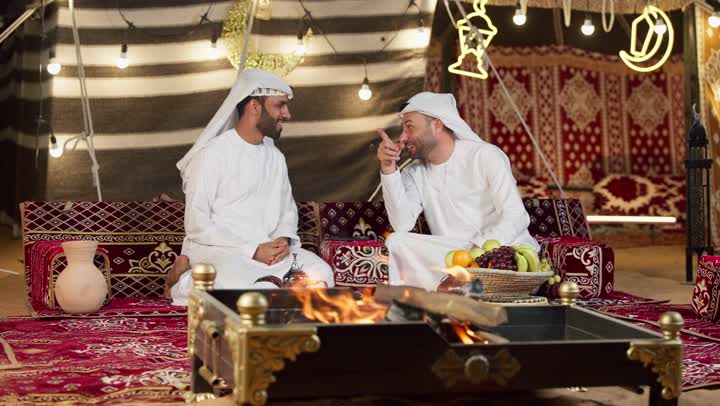 Traditional Majlis, two smiling female friends sitting in front of a firewood bonfire, two Arab Gulf men and two females talking about something, a gesture pointing to something, night camping in the UAE desert, generous hospitality and warm welcome