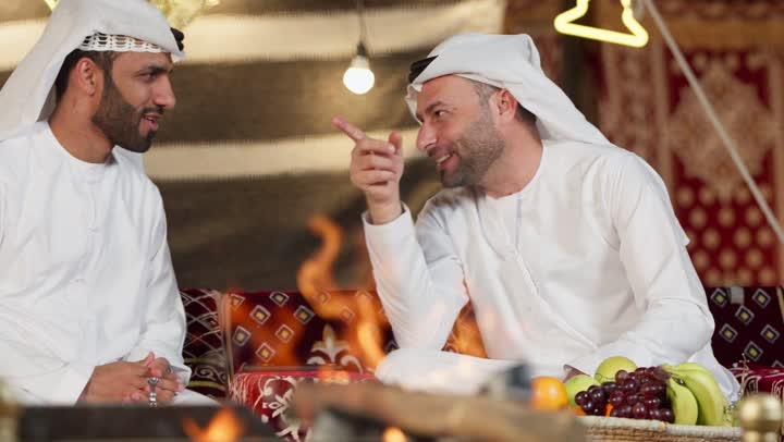 عادات وتقاليد المجلس الشعبي، رجلان عربيان خليجيان اماراتيان يجلسان امام شعله الحطب، كرم وحسن الضيافه العربيه، شاب اماراتي يشير الي شئ ما، جلسه وكشته برشبابيه