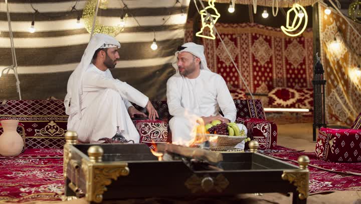 Traditional People's Council, two smiling female friends sitting in front of a firewood bonfire, two Arab Gulf men and two females talking about something, night camping in the UAE desert, generous hospitality and warm welcome