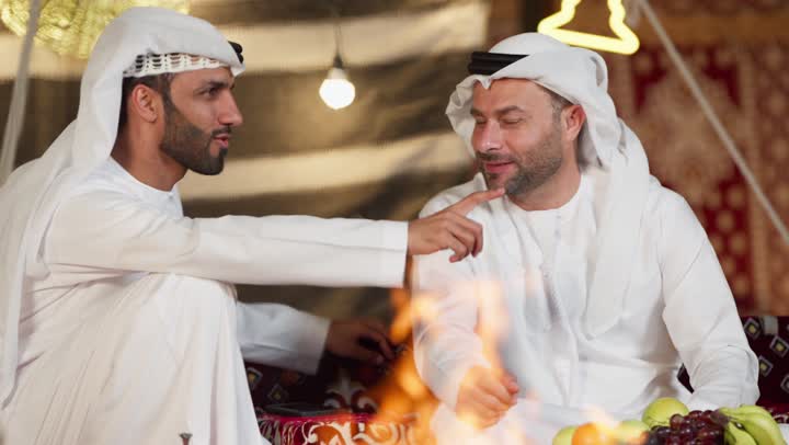 ايماءه الاشاره الي شئ ما، المجلس الشعبي التقليدي، صديقان امراتيان مبتسمان يجلسان امام شعله الحطب، رجلان عربيان خليجيان امراتيان يتحدثان عن امر ما، التخييم الليلي في صحراء الامارات، كرم الضيافه وحسن الاستقبال