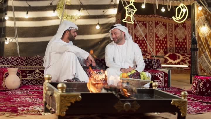 رجلان عربيان خليجيان اماراتيان يتحدثان يجلسان امام شعله الحطب، عادات وتقاليد المجلس الشعبي، كرم وحسن الضيافه العربيه، شاب اماراتي يشير الي شئ ما، جلسه وكشته برشبابيه