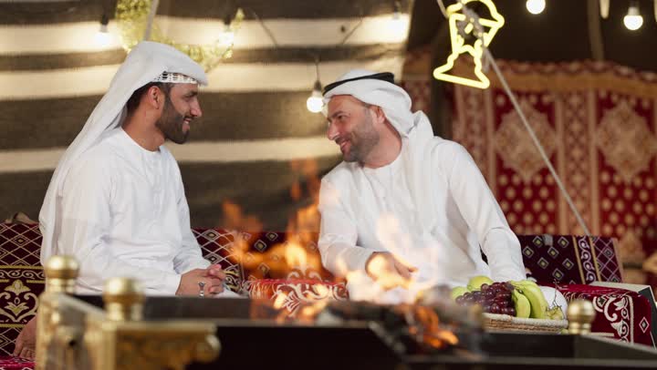 رجلان عربيان خليجيان اماراتيان يتحدثان يجلسان امام شعله الحطب، عادات وتقاليد المجلس الشعبي، كرم وحسن الضيافه العربيه، شاب اماراتي يشير الي شئ ما، جلسه وكشته برشبابيه