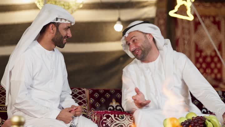 صديقان امراتيان مبتسمان يجلسان امام شعله الحطب، رجل عربي خليجي اماراتي يقدم لصديقه علبه بمناسبه شهر الكرم، التخييم الليلي في صحراء الامارات، كرم الضيافه وحسن الاستقبال، المجلس الشعبي التقليدي، خيمه مليئه بالزينه والديكور الرمضاني