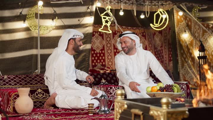 صديقان امراتيان مبتسمان يجلسان امام شعله الحطب، رجل عربي خليجي اماراتي يقدم لصديقه علبه بمناسبه شهر الكرم، التخييم الليلي في صحراء الامارات، كرم الضيافه وحسن الاستقبال، المجلس الشعبي التقليدي، خيمه مليئه بالزينه والديكور الرمضاني