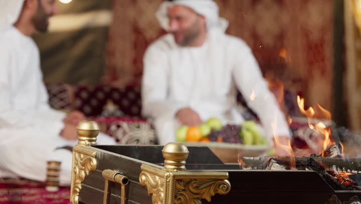تصوير مقرب لشعله الحطب في أجواء ليلية هادئة، رجلان عربيان خليجيان اماراتيان يتحدثان يجلسان في خيمه بدويه عربيه، عادات وتقاليد المجلس الشعبي، كرم وحسن الضيافه العربيه، شاب سعودي يتحدث مع صديقه