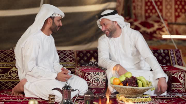 كرم الضيافه وحسن الاستقبال، صديقان امراتيان مبتسمان يجلسان امام شعله الحطب، رجل عربي خليجي اماراتي يقدم لصديقه بعض الفواكهه، التخييم الليلي في صحراء الامارات، المجلس الشعبي التقليدي، خيمه مليئه بالزينه والديكور الرمضاني