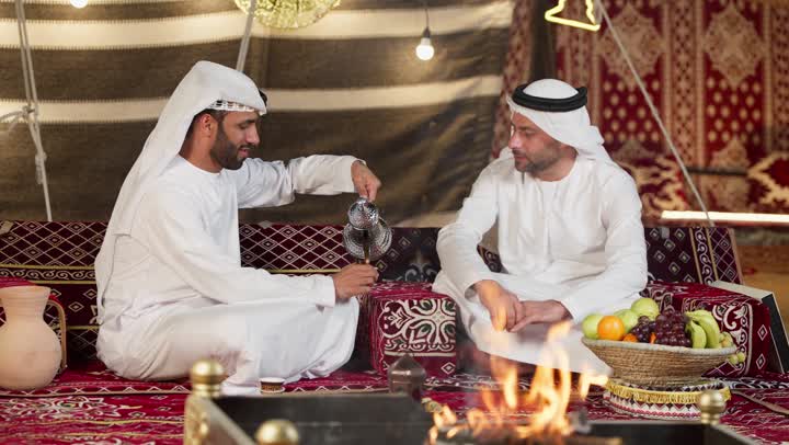 عادات وتقاليد أصيلة في تقديم المشروبات, السير على نهج الأجداد في إكرام الضيف,  رجل عربي خليجي اماراتي يرتدي الكندوره والغتره يقوم بصب القهوة من الدلة النحاسية لصديقه, توارث حسن الاستقبال والكرم, تقديم المنتجات المحلية للضيوف،المجلس الشعبي في الامارات