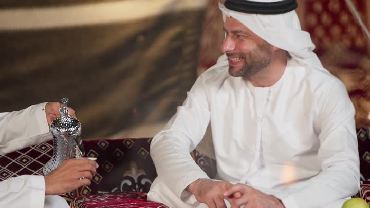 Good reception for friends in the People's Council, a close-up of a young Emirati Gulf Arab man wearing a kandura and ghutra drinking hot coffee, chatting and talking about something, a close-up of a young man holding a copper coffee pot and offering his friend coffee, honoring the guest and good reception